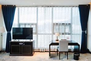 A hotel room with a large flat-screen TV on a dark wooden cabinet, a desk with a chair, and a large window with sheer curtains. - Tianrui Hotel (Yanhe)