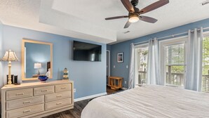 Un dormitorio con cama, cómoda, televisor, ventilador de techo y ventanas amplias.