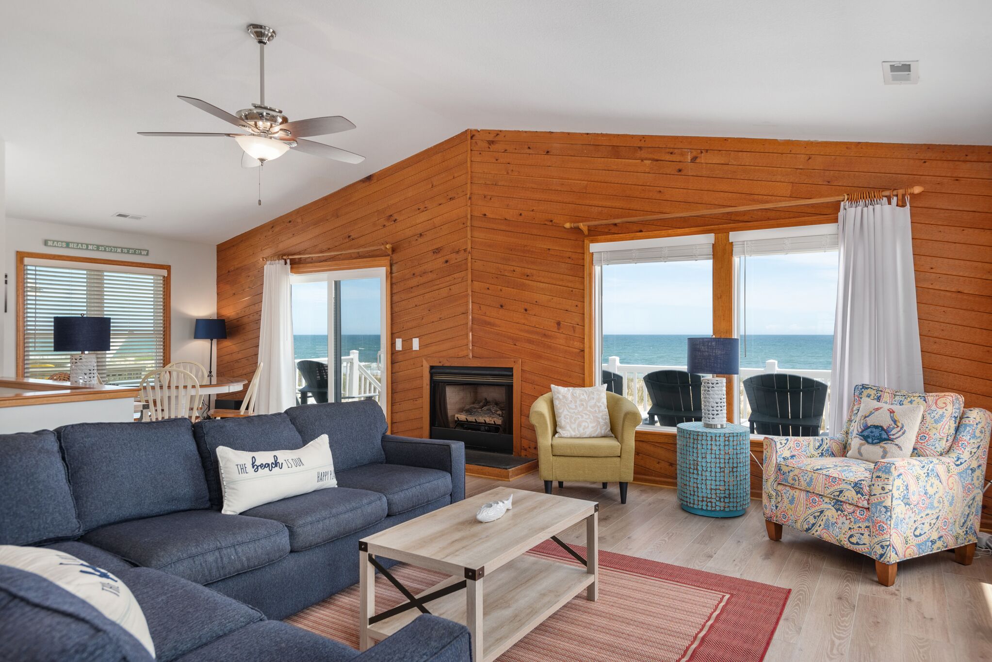 A living room with a sofa, armchairs, a coffee table, and a fireplace. Large windows offer a view of the sea.