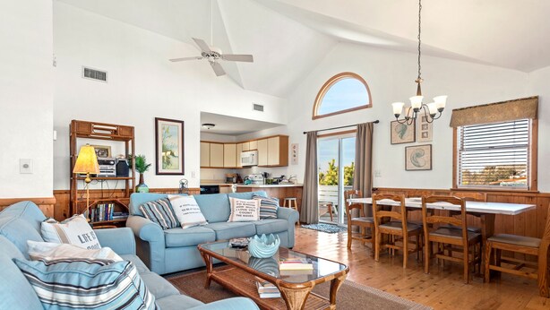 A spacious living room with a blue sofa, a glass coffee table, and a dining area with wooden chairs and a table. - Kiki's Retreat: Walk to Duck Village | Pets OK (Duck)