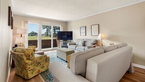 Un salon comprenant un canapé, un fauteuil et un pouf, un téléviseur à écran plat et une grande fenêtre donnant sur un paysage verdoyant.