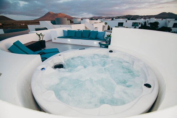 A hot tub surrounded by white furniture with blue cushions, set against a backdrop of a mountainous landscape.
