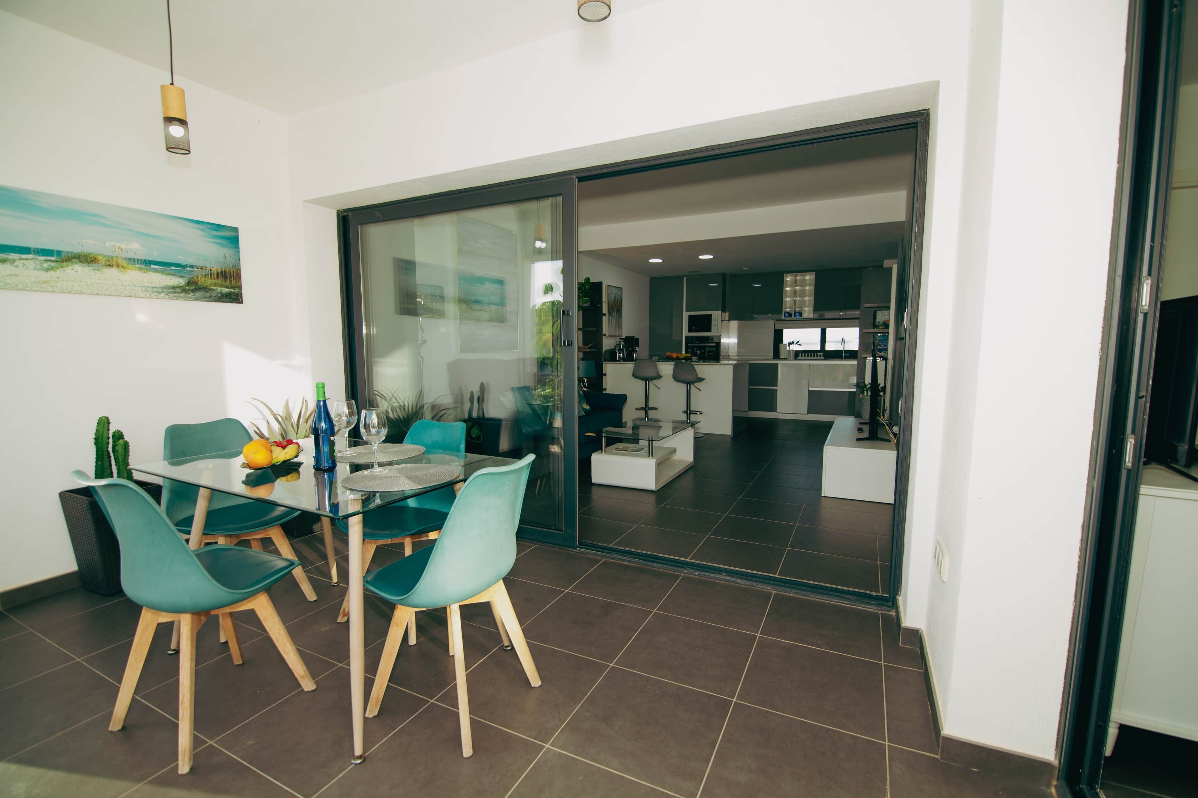Un comedor moderno con una mesa y sillas de vidrio, una cocina al fondo y un piso de vidrio corredizo que da a otra habitación.