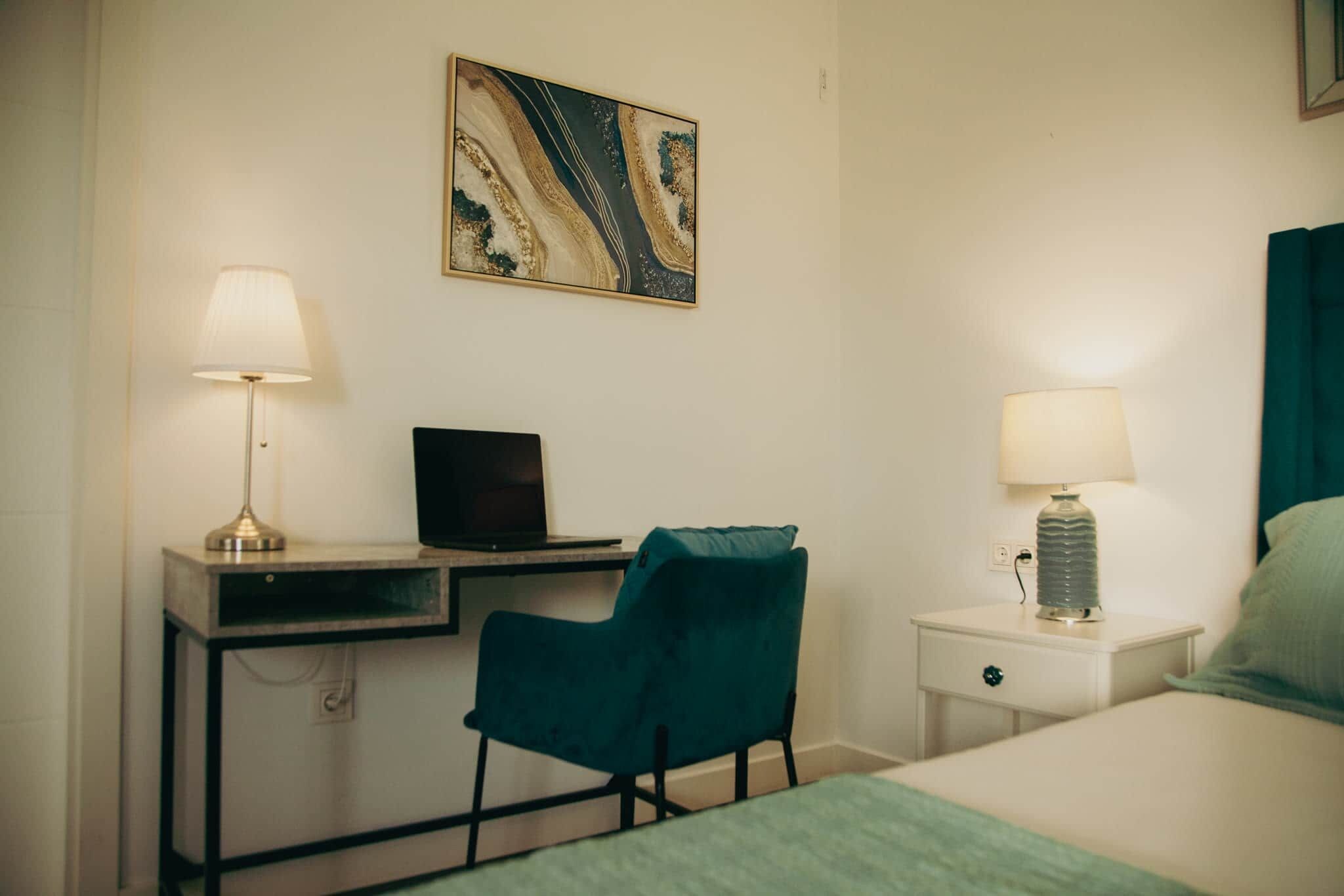 A hotel room with a desk, chair, bed, bedside table, lamps, and a wall-mounted artwork.