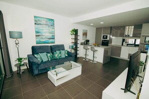 A modern living room with a sofa, coffee table, and a kitchen area with a bar and appliances.