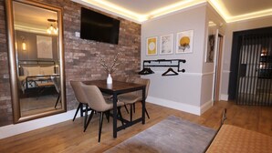 A modern living room with a brick wall, a large mirror, a dining table with chairs, a TV mounted on the wall, and a coat rack with hangers. - Alma Studios - The Safe Room - Hopewell (Bristol)