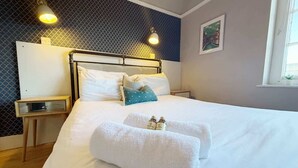 Una cama bien hecha con sábanas blancas y dos toallas dobladas, una mesita de noche con una lámpara, y una ventana con vistas al exterior.