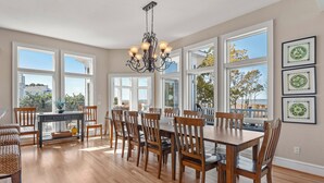 A spacious dining area with a large wooden table and chairs, a chandelier, and large windows offering a view of the outdoors.