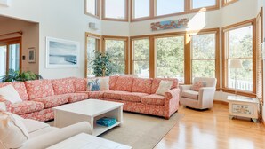 Un salon spacieux avec un grand canapé d’angle, une table basse et une table d’appoint. La pièce est dotée de grandes fenêtres à cadre en bois et offre une vue sur la verdure environnante.