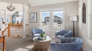 A spacious living room with a high ceiling, a chandelier, wicker coffee table, blue patterned chairs, and large windows offering a view of the outside. - It's 5 O'clock Somewhere: Pool, Spa, Club Access (Corolla)