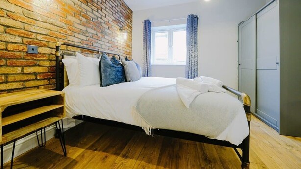 A bedroom with a brick wall, a bed with white bedding, a wooden bench, a window with curtains, and a wardrobe. - One Bedroom Apartment Close to MOD (Bristol)
