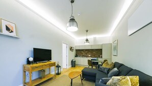 A modern living room with a dark sofa, wooden console table, and a flat-screen TV. A kitchen with a brick backsplash and white cabinets is visible in the background. - One Bedroom Apartment Close to Cribbs Causeway! (Bristol)