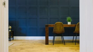 Um quarto com parede revestida em azul escuro, uma mesa de madeira com duas cadeiras, e uma pequena planta sobre a mesa.