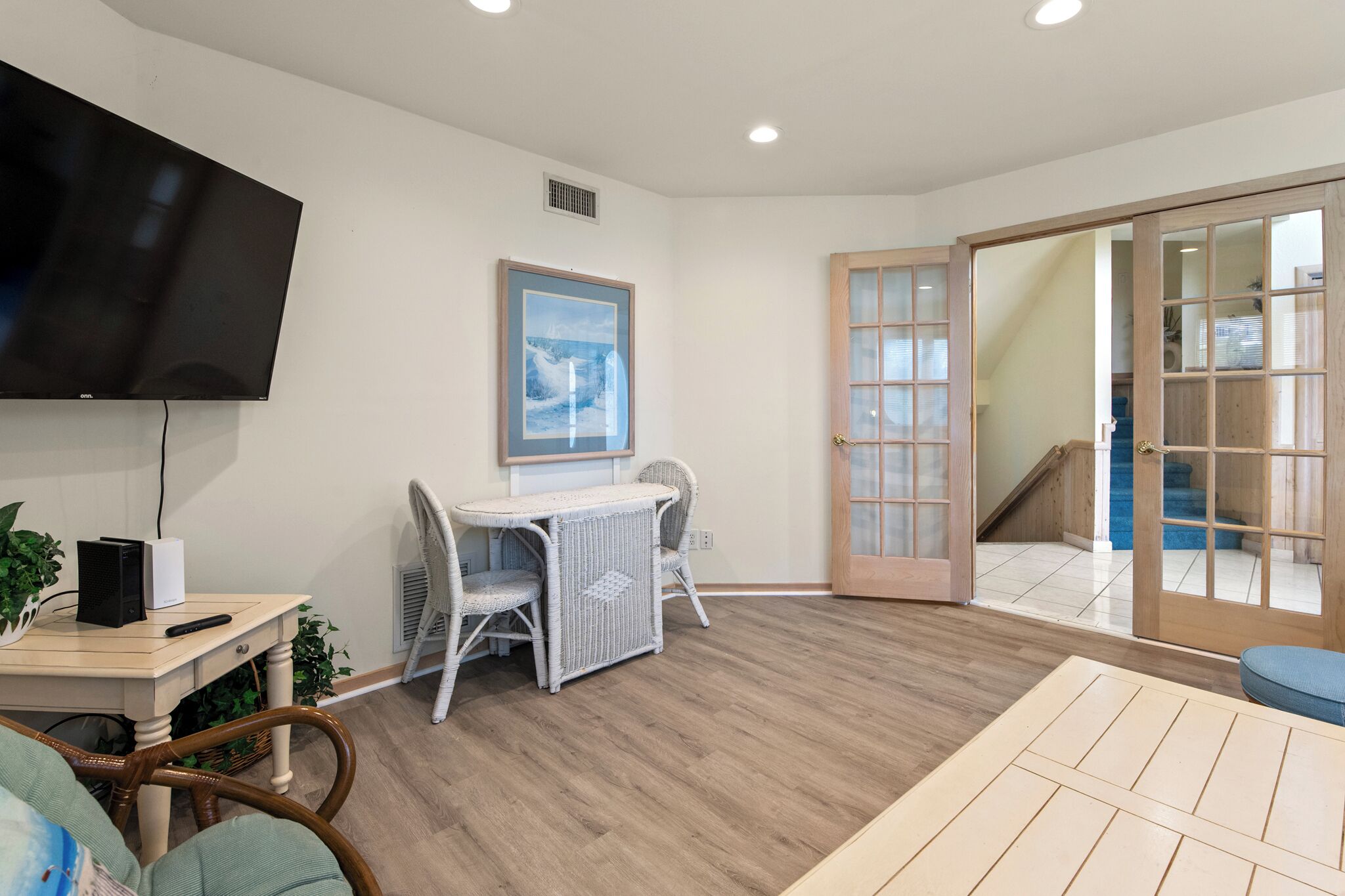 A room with a flat-screen TV, a wicker table, and chairs. There is a wooden door with glass panels leading to a staircase.