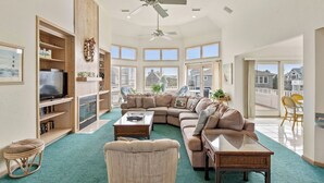 A spacious living room with a sectional sofa, a television, and a large window offering a view of the neighborhood. - Payson Place: Oceanside, Pool, Golf Discounts, Spa (Corolla)