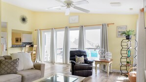 Una sala de estar con un sofá, una mesa de centro y un sillón reclinable. Hay un ventilador de techo, una ventana con cortinas y vistas al exterior.