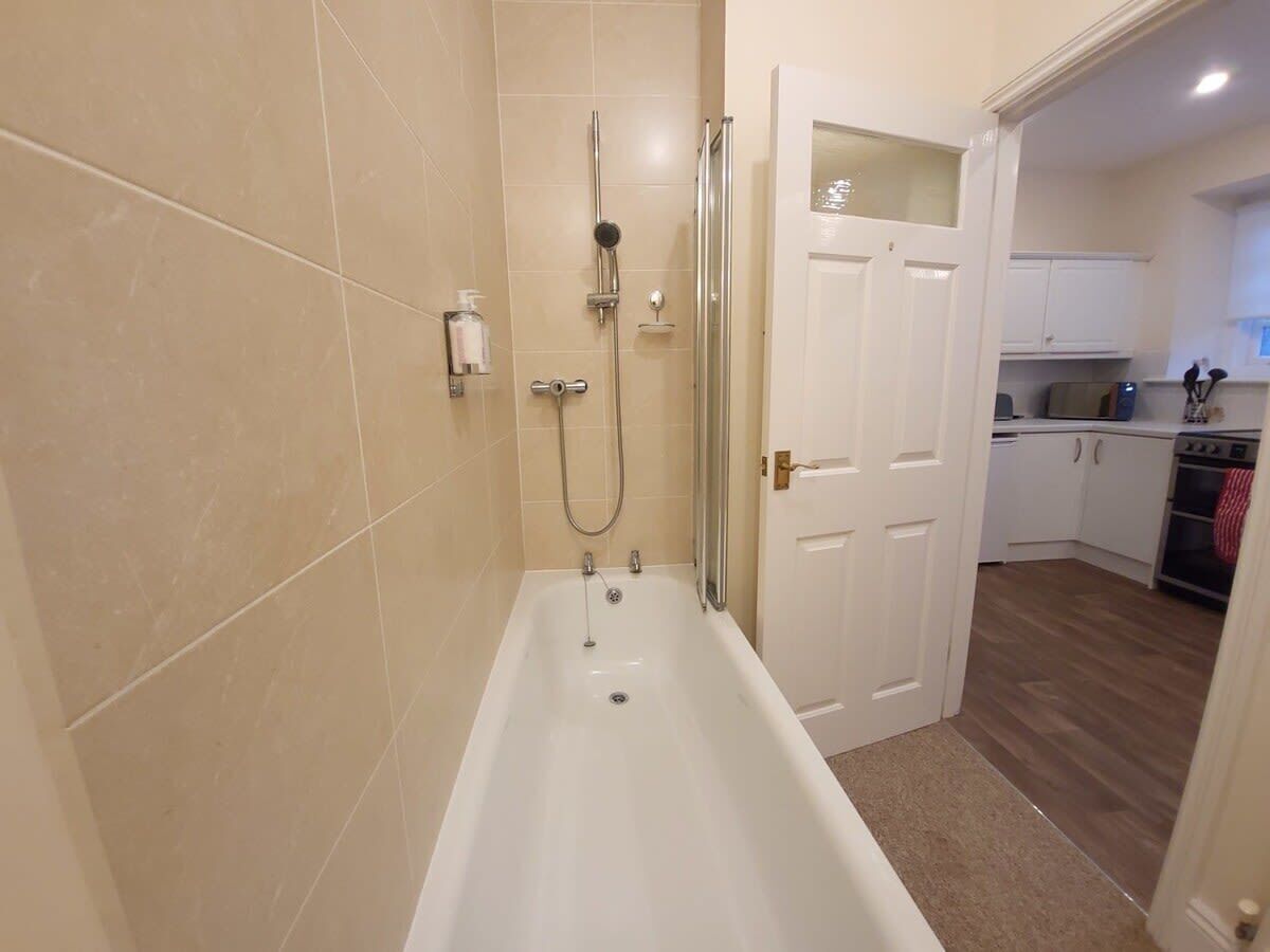 Un baño con bañera, ducha e inodoro. Se ve una cocina al fondo.