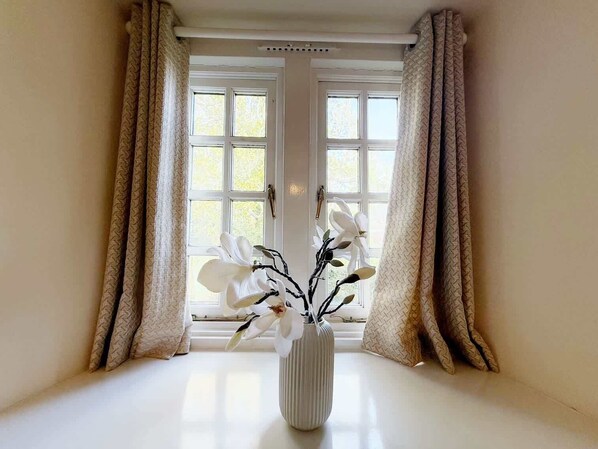 A room with a large window, white curtains, and a vase with white flowers on a white table.