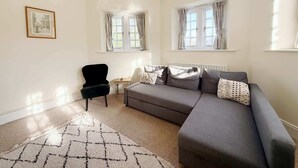 Ein modernes Wohnzimmer mit einem grauen Sofa, einem schwarzen Sessel und einem gemusterten Teppich. Zwei Fenster mit Vorhängen, ein Heizkörper und ein gerahmtes Bild sind zu sehen.