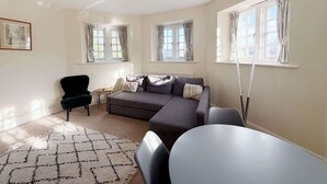 Ein modernes Wohnzimmer mit einem grauen Sofa, einem schwarzen Sessel, einem runden Tisch und einem gemusterten Teppich. Drei Fenster mit Vorhängen, ein Heizkörper unter dem Sofa und eine Deckenleuchte sind zu sehen.