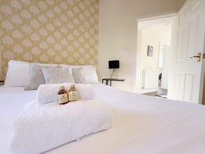 A neatly made bed with white linens and a fluffy white towel, two bottles of ELSYL, a black lamp, and a patterned wallpaper.