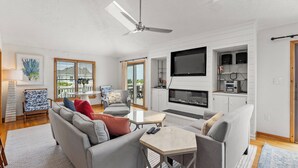 A modern living room with a sofa, coffee table, TV, fireplace, and a ceiling fan. - Sea La Vie: Oceanside, Hot Tub, Shared Pool (Corolla)