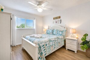 Une chambre à coucher comprenant un lit, des tables de chevet, un ventilateur de plafond, une fenêtre avec des stores et un panneau décoratif mural.