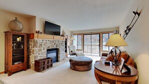 A living room with a stone fireplace, a flat-screen TV mounted above it, a comfortable armchair, a round wooden table with a lamp, and a large window offering a view of the outside. - Boutique Complex Steps Away from Mammoth! Hike, Mountain Bike, Free WiFi and Free Parking! by RedAwning (Mammoth Lakes)