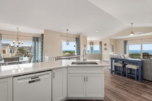 A modern kitchen with white cabinetry, a Bosch dishwasher, and a large island with a sink. There is a dining area with a blue table and chairs, and a view of the ocean through large windows. - Dune Ridge: Oceanfront Duplex, Spa, YMCA Access (Nags Head)