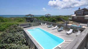 Una terraza de madera con piscina, sillas de descanso y quioscos con vista al océano.