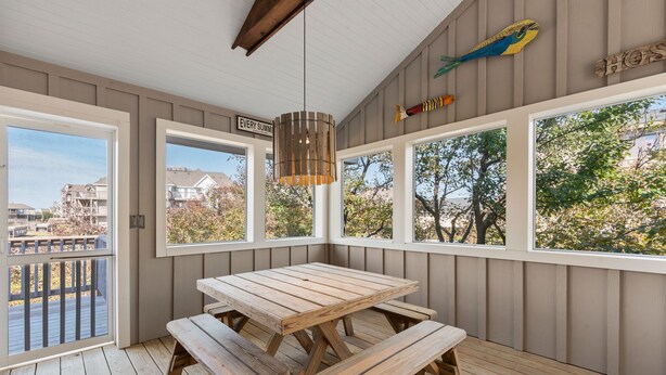 Une terrasse en bois avec une table de pique-nique et des bancs, de grandes fenêtres offrant une vue sur les arbres et les bâtiments, et un luminaire suspendu.