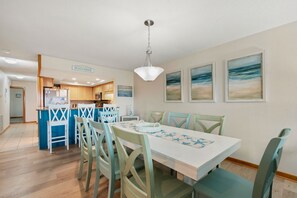 A dining area with a white table and teal chairs, a kitchen in the background, and a beach-themed wall art.
