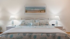 Een slaapkamer met een bed, twee nachtkastjes, een lamp, een kussen met de tekst 'Seaside' en een lijst met een strandlandschap.