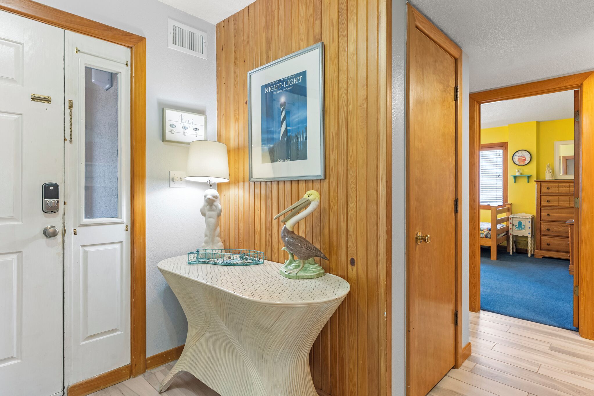 A hallway with a wooden door, a wall-mounted lamp, a framed poster, and a decorative pelican sculpture.