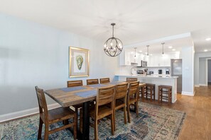 A modern kitchen with white cabinets, a central island, and a dining area with wooden chairs and a table. - Heron Cove 302N: Oceanfront Condo, Top Floor, Pool (Nags Head)