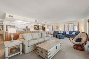 Ein modernes Wohnzimmer mit Sofa, Sessel und Couchtisch, im Hintergrund eine Küche und ein Essbereich mit Tisch und Stühlen.