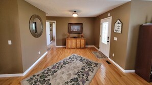 Un hall spacieux doté d’un parquet en bois, d’un grand tapis à motifs floraux, d’une console en bois avec un miroir et d’un tableau encadré accroché au mur.