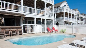 A pool area with a two-story building, outdoor seating, and a clear blue sky. - On Holiday: Oceanside, Private Pool, Spa (Nags Head)