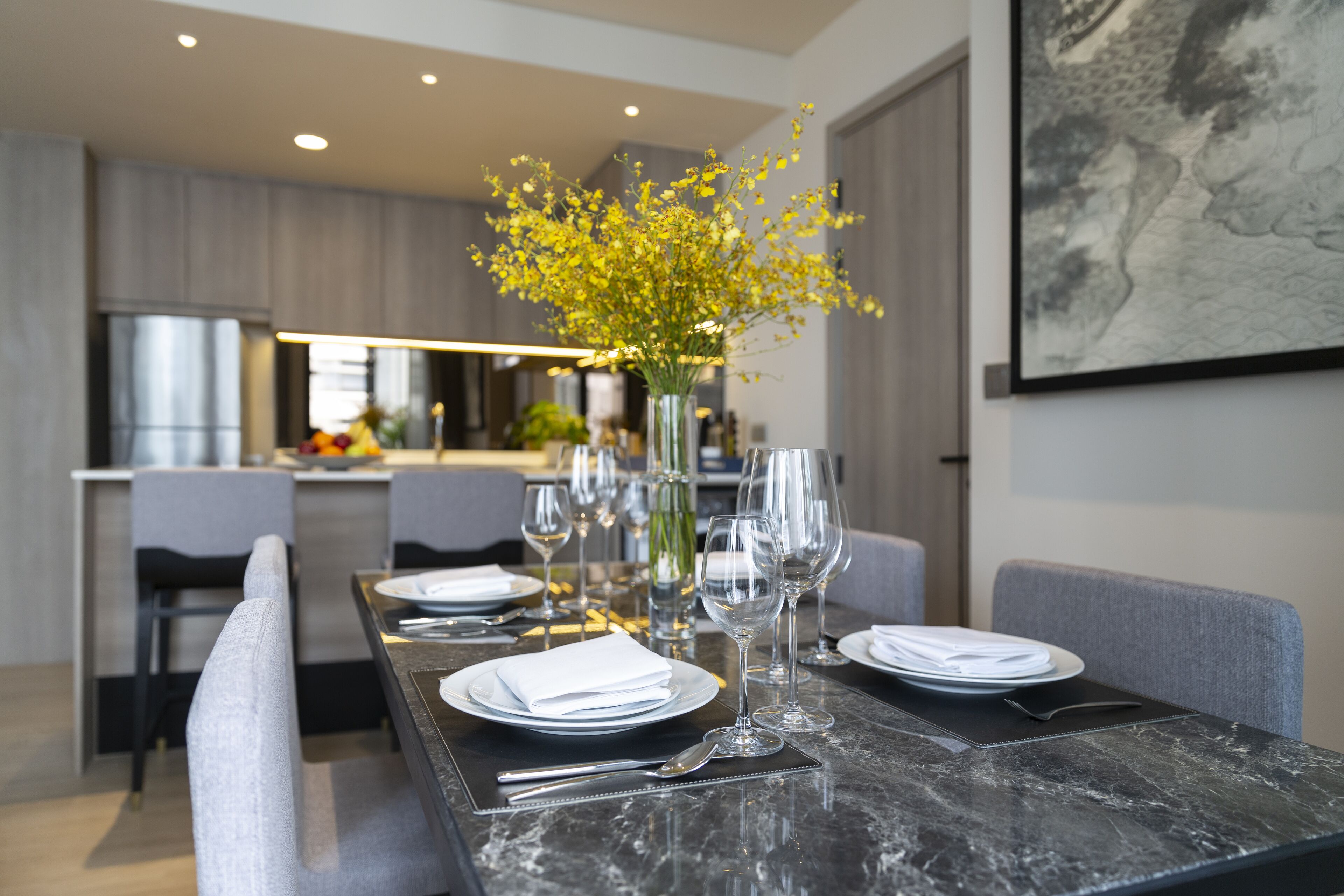A modern dining area with a marble table set for a meal, a vase with yellow flowers, and a large abstract painting on the wall.