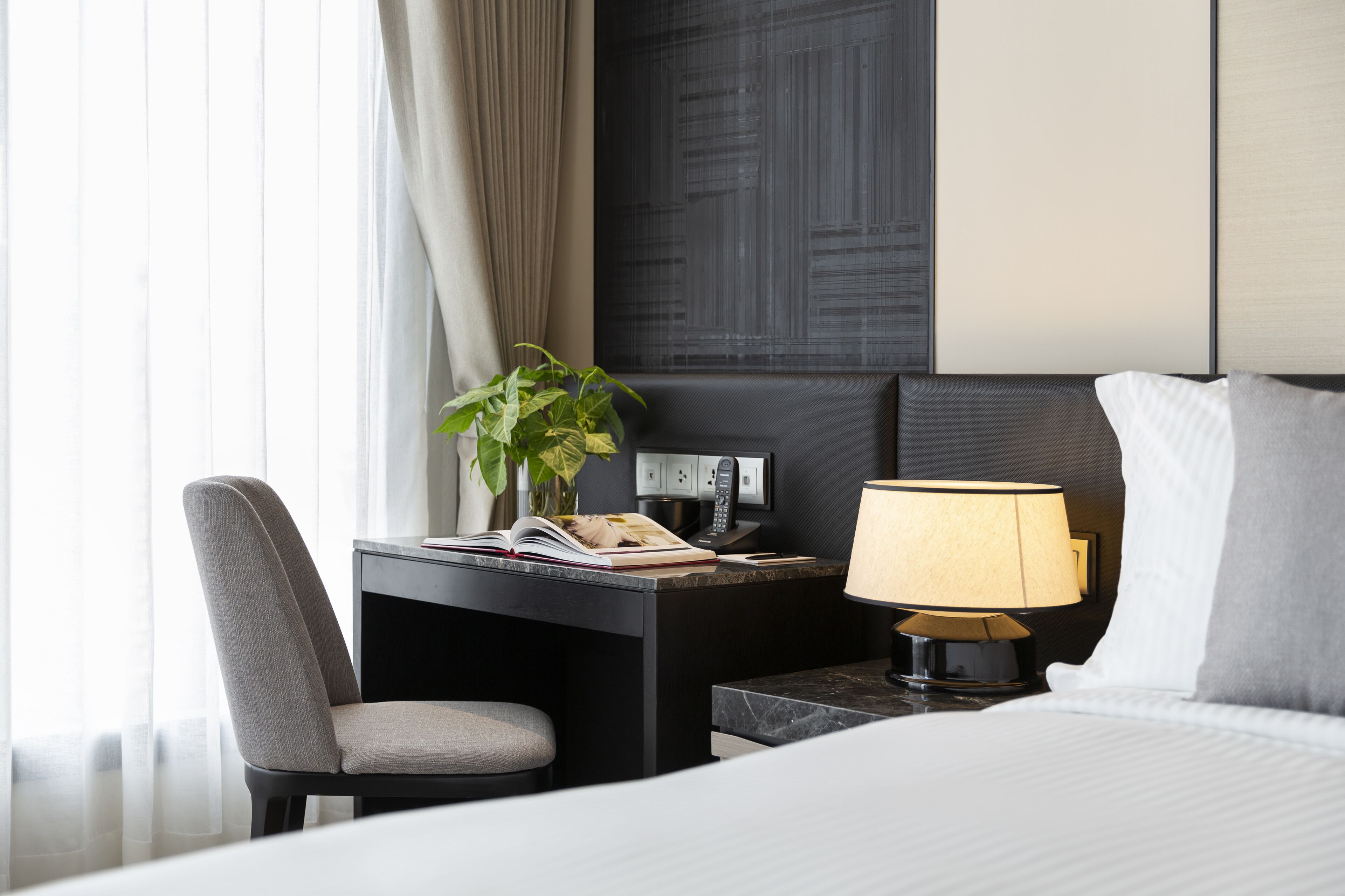 A hotel room with a bed, a desk with a lamp, a chair, and a plant.