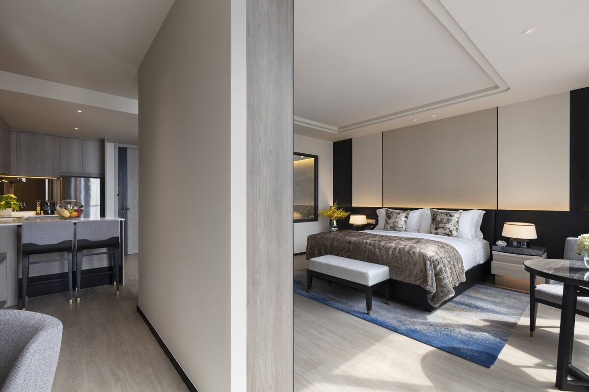 A modern hotel room with a bed, bedside table, lamp, and a small desk area with a chair.