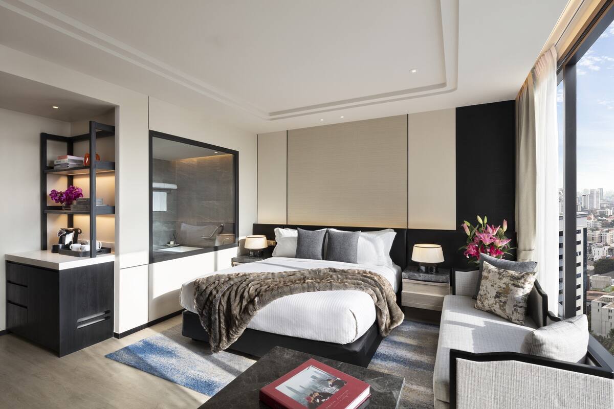 A modern hotel room with a large bed, a sitting area with a sofa and armchair, a desk with a chair, a large window with a city view, and a built-in bookshelf.