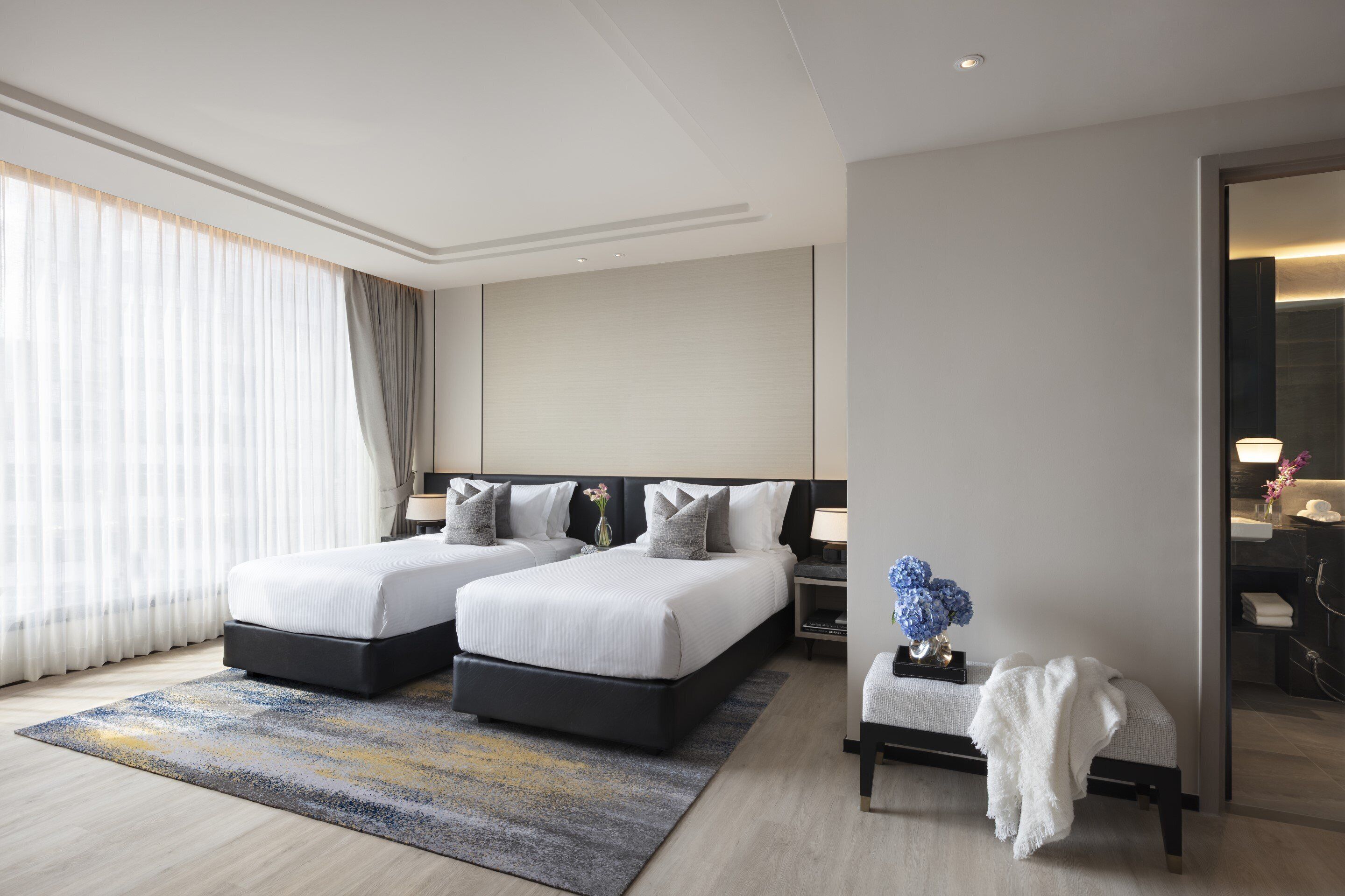 Une chambre d’hôtel moderne avec deux lits, un coin salon et une salle de bain visible à travers une porte ouverte.