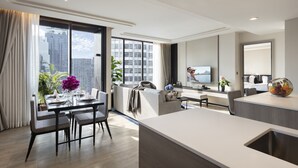 A modern hotel room with a dining area, a sofa, a TV, and a large window with a city view.