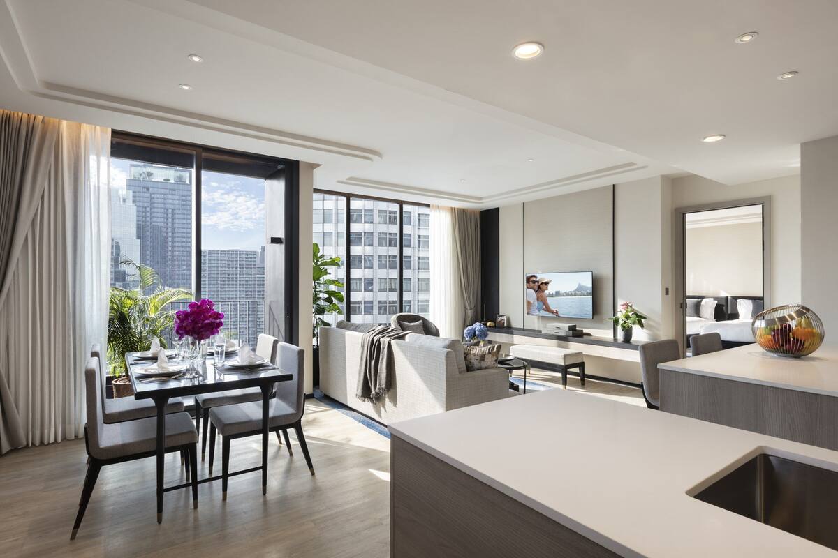 A modern hotel room with a dining area, a sofa, a TV, and a large window with a city view.