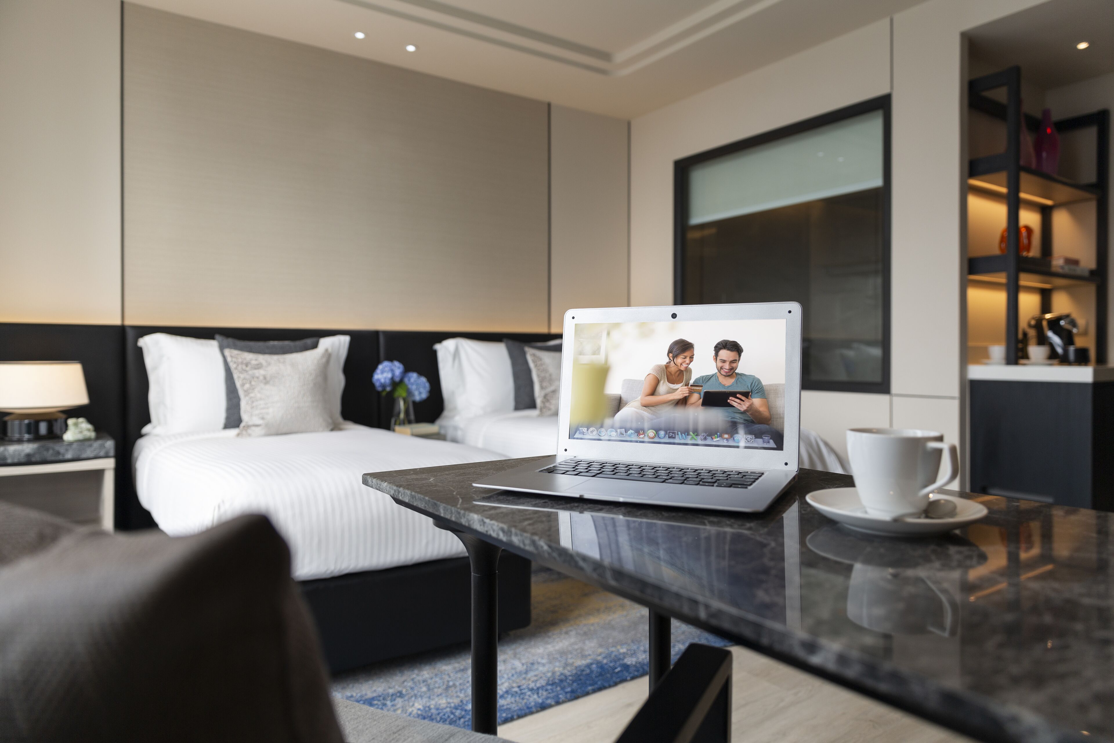 Une chambre d’hôtel moderne avec un lit, un ordinateur portable posé sur une table, une tasse et une vue sur le paysage urbain par la fenêtre.