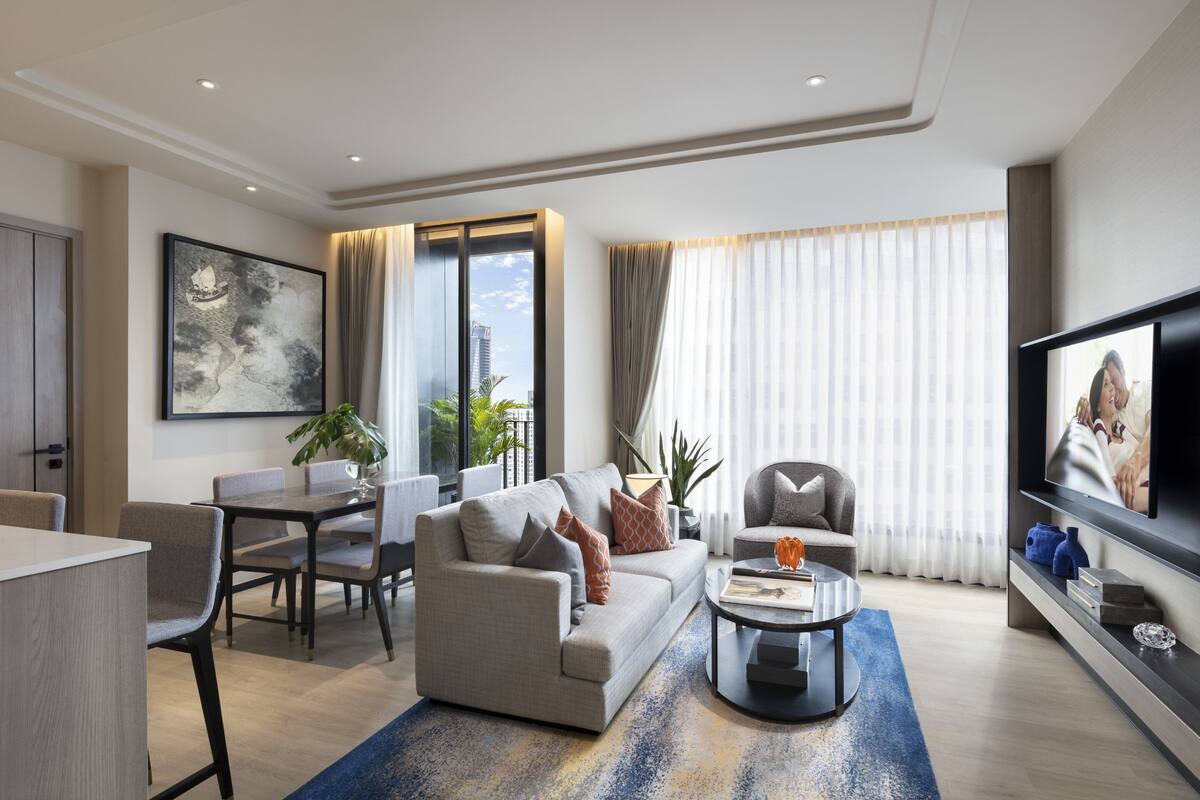 A modern living room with a sofa, coffee table, and TV. There is a dining area with chairs and a table. The room has large windows with curtains.