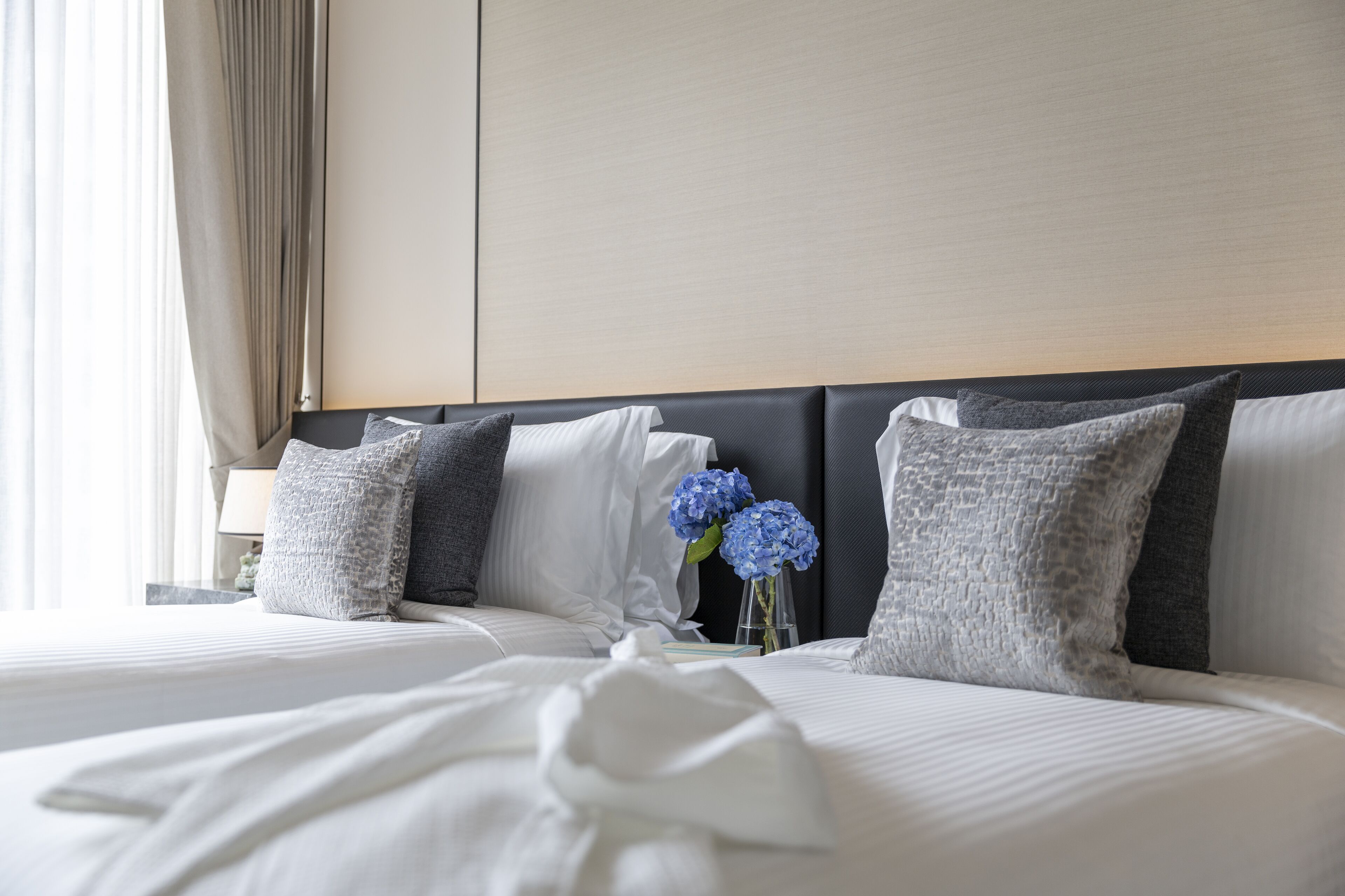 Una cama bien tendida con sábanas blancas y almohadas grises, un florero con flores azules y un cabecero moderno.
