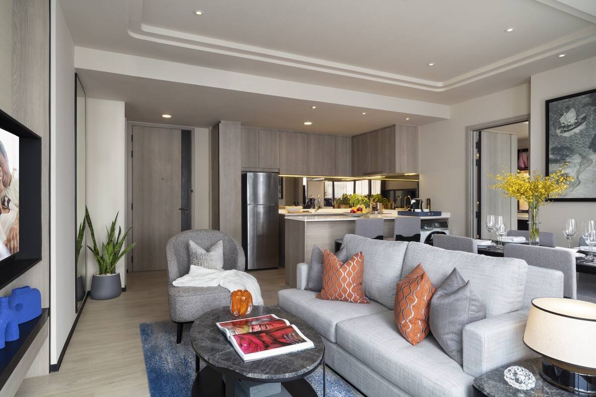 A modern living room with a grey sofa, a coffee table, and a TV on a stand. There is a kitchen area with a fridge and a dining table with chairs.