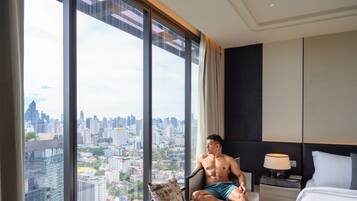 A hotel room with a large window offering a city view, a sofa, a bedside table with a lamp, and a bed.